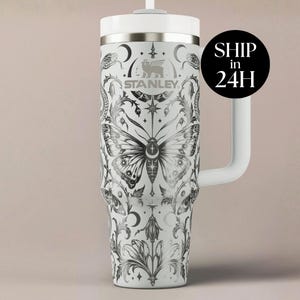May include: A white Stanley tumbler with a handle and a straw, featuring a detailed black graphic print of a large central moth, snakes, crescent moons, stars, and intricate floral scrollwork patterns covering the entire exterior surface.