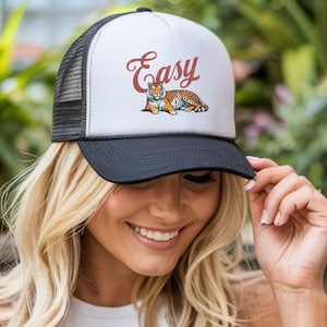 May include: A black and white trucker hat featuring the word "Easy" in cursive and a tiger illustration. The hat has a mesh back and a curved peak. The hat is worn by a person.