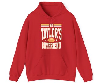 Peut inclure: Un sweat-shirt à capuche rouge avec une poche avant et un cordon de serrage. Le devant affiche le texte "60 TAYLOR'S BOYFRIEND" en blanc, dans une police de style universitaire, avec un ballon de football.