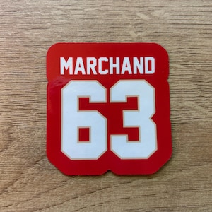 Puede incluir: Una pegatina cuadrada roja con el nombre "MARCHAND" en letras blancas en bloque sobre el n&uacute;mero "63" en blanco con un contorno rojo. La pegatina est&aacute; sobre una superficie de madera.
