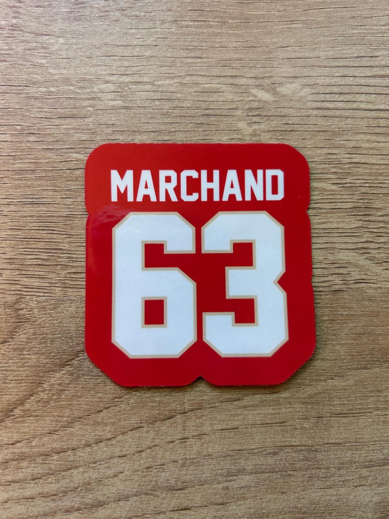 Puede incluir: Una pegatina cuadrada roja con el nombre "MARCHAND" en letras blancas en bloque sobre el n&uacute;mero "63" en blanco con un contorno rojo. La pegatina est&aacute; sobre una superficie de madera.