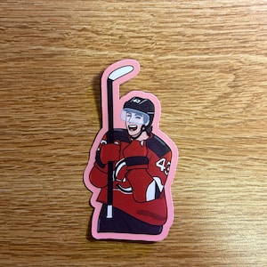May include: A sticker featuring a cartoon hockey player in a red jersey with the number 43. The player is wearing a black helmet and holding a hockey stick. The sticker has a pink border and is on a wooden surface.