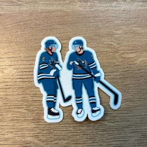 Puede incluir: Dos pegatinas de jugadores de hockey con uniformes azules y blancos, cascos y palos de hockey. Un jugador tiene el número 2 en su camiseta, el otro el número 7. Las pegatinas están sobre una superficie de madera.