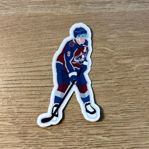 May include: A sticker featuring a hockey player in a blue, burgundy, and white uniform, with the number 8 visible. The player is in a skating pose, holding a hockey stick. The sticker has a white border.