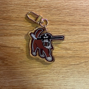 May include: A clear acrylic keyring featuring a cartoon dog in brown and white, holding a long, black object. The keyring has a gold-coloured clasp. The dog is outlined in a dark red colour.