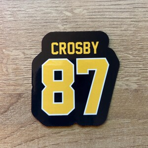 Puede incluir: Una pegatina negra y dorada con el nombre "CROSBY" sobre el n&uacute;mero "87". El n&uacute;mero est&aacute; en negrita, fuente amarilla con un contorno negro. La pegatina tiene forma rectangular redondeada.