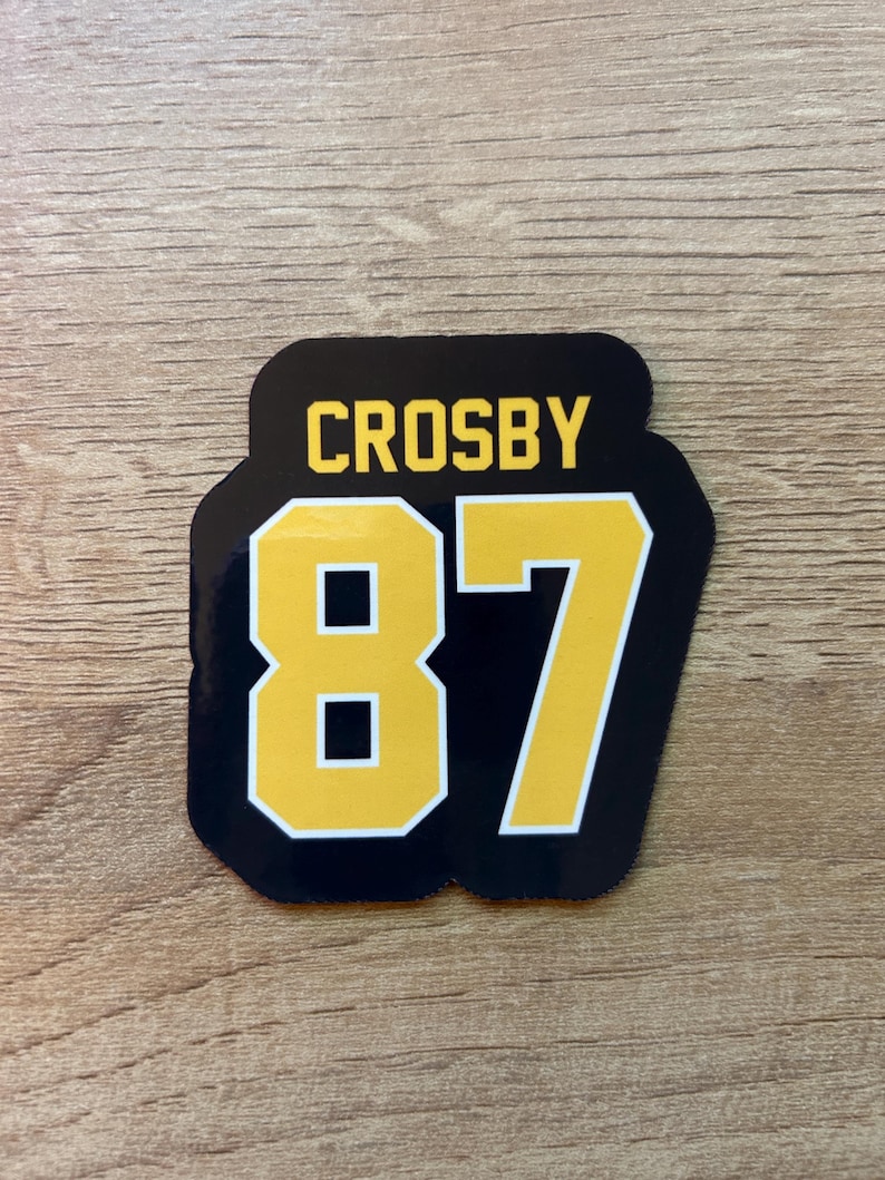 Puede incluir: Una pegatina negra y dorada con el nombre "CROSBY" sobre el n&uacute;mero "87". El n&uacute;mero est&aacute; en negrita, fuente amarilla con un contorno negro. La pegatina tiene forma rectangular redondeada.