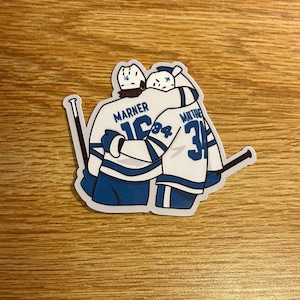 May include: A sticker featuring two hockey players in white and blue jerseys embracing. The jersey on the left reads "Marner 16" and the jersey on the right reads "Matthews 34". Hockey sticks are visible on either side of the players.