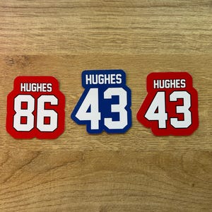 Puede incluir: Tres pegatinas rectangulares con el nombre "HUGHES" encima de n&uacute;meros grandes. Una pegatina es roja con texto blanco y el n&uacute;mero "86". Las otras dos pegatinas son azules y rojas con el n&uacute;mero "43".