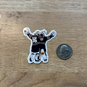 Puede incluir: Una pegatina de un portero de hockey con uniforme negro y dorado, con el n&uacute;mero 35. El portero est&aacute; representado con los brazos levantados. Una moneda est&aacute; al lado de la pegatina para comparar el tama&ntilde;o.