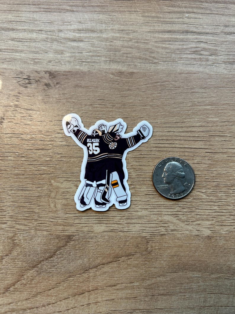 Puede incluir: Una pegatina de un portero de hockey con uniforme negro y dorado, con el n&uacute;mero 35. El portero est&aacute; representado con los brazos levantados. Una moneda est&aacute; al lado de la pegatina para comparar el tama&ntilde;o.