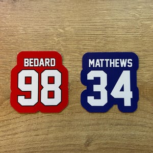 Puede incluir: Dos pegatinas estilo camiseta. Una es roja con ribete blanco, el nombre "BEDARD" encima del n&uacute;mero "98". La otra es azul con ribete blanco, el nombre "MATTHEWS" encima del n&uacute;mero "34".