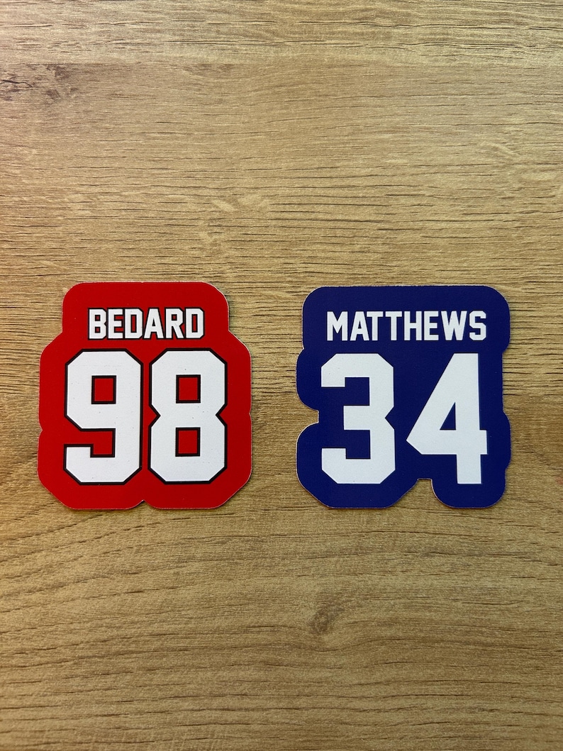 Puede incluir: Dos pegatinas estilo camiseta. Una es roja con ribete blanco, el nombre "BEDARD" encima del n&uacute;mero "98". La otra es azul con ribete blanco, el nombre "MATTHEWS" encima del n&uacute;mero "34".