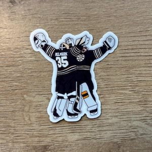 Può includere: Un adesivo con due portieri di hockey in uniforme nera e oro, con le braccia alzate in segno di festa. Il numero 35 e il nome "ULLMARK" sono stampati sul retro di una maglia. È visibile anche il logo dei Boston Bruins.