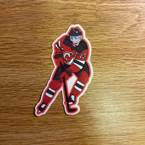Puede incluir: Una pegatina con un jugador de hockey de dibujos animados con uniforme rojo y negro con el número 13. El jugador lleva casco y sostiene un palo de hockey. La pegatina tiene un borde blanco.