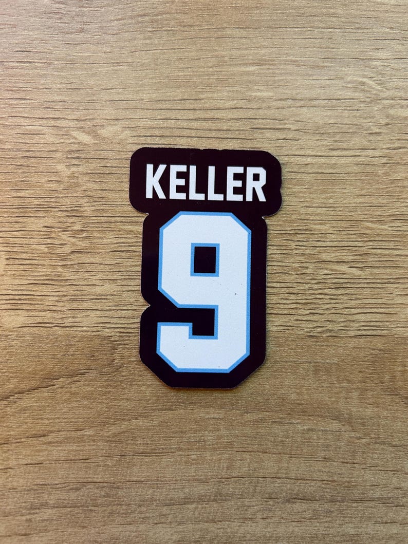 Puede incluir: Una pegatina con el nombre "KELLER" encima del n&uacute;mero "9". El n&uacute;mero es blanco con un contorno azul claro y un interior azul oscuro. El nombre "KELLER" est&aacute; en may&uacute;sculas blancas sobre un fondo oscuro.