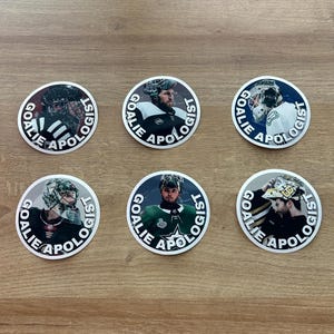 May include: Six circular stickers with images of hockey goalies. Each sticker displays a different goalie and the text "GOALIE APOLOGIST" in a circular arrangement. The stickers are presented on a light brown surface.
