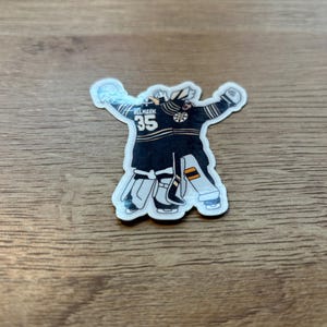 Puede incluir: Una pegatina de un portero de hockey con uniforme negro y dorado, con el n&uacute;mero 35. El portero est&aacute; representado con los brazos levantados. La pegatina tiene un borde blanco y est&aacute; sobre una superficie de madera.