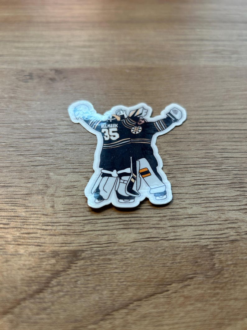 Puede incluir: Una pegatina de un portero de hockey con uniforme negro y dorado, con el n&uacute;mero 35. El portero est&aacute; representado con los brazos levantados. La pegatina tiene un borde blanco y est&aacute; sobre una superficie de madera.