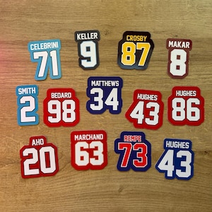 May include: A collection of colorful hockey jersey-style number stickers. Each sticker features a player's name and number, with various color schemes including blue, red, and yellow. The numbers range from 2 to 98.