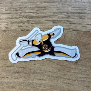 Puede incluir: Una pegatina que representa a un portero de hockey en una pose de buceo. El portero lleva un uniforme negro, dorado y blanco con el logotipo de los Bruins de Boston. La pegatina tiene un borde blanco y está sobre una superficie de madera.