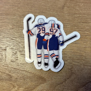 May include: A sticker featuring two hockey players in blue and orange uniforms, embracing. One player has the number 29, the other 97. Both hold hockey sticks. The sticker has a white border and is on a wooden surface.
