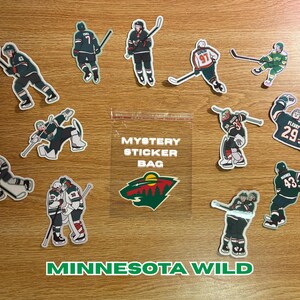 Pode incluir: Uma coleção de autocolantes de hóquei do Minnesota Wild e um "Mystery Sticker Bag" numa superfície de madeira. Os autocolantes apresentam jogadores com uniformes verdes, brancos e pretos. O logótipo do Minnesota Wild também é visível.