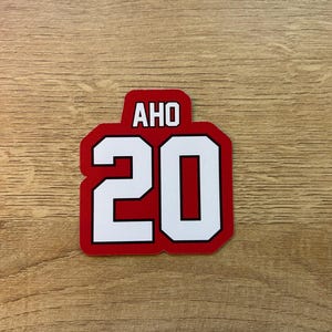 Puede incluir: Una pegatina roja y blanca con el texto "AHO" encima del n&uacute;mero "20". Los n&uacute;meros y letras blancos tienen un contorno negro sobre un fondo rojo. La pegatina es rectangular con esquinas redondeadas.
