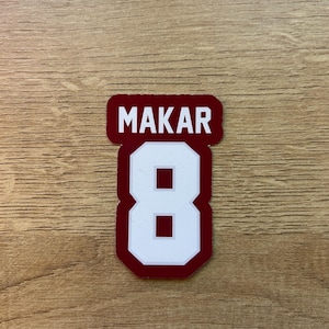 Puede incluir: Una pegatina granate y blanca con el nombre "MAKAR" encima del n&uacute;mero "8". El n&uacute;mero es blanco con un contorno granate. La pegatina est&aacute; sobre una superficie de madera.