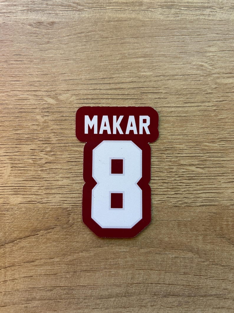 Puede incluir: Una pegatina granate y blanca con el nombre "MAKAR" encima del n&uacute;mero "8". El n&uacute;mero es blanco con un contorno granate. La pegatina est&aacute; sobre una superficie de madera.