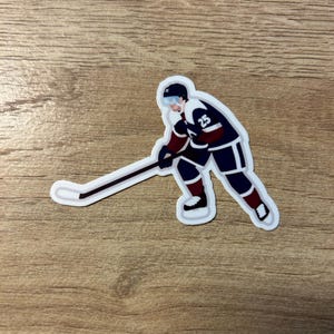 Könnte beinhalten: Aufkleber mit einem Eishockeyspieler in einem marineblauen und weinroten Trikot mit der Nummer 25. Der Spieler ist in einer Skating-Pose und hält einen Hockeyschläger. Der Aufkleber hat einen weißen Rand.