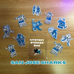 Peut inclure: Divers autocollants des Sharks de San José, représentant des joueurs de hockey en bleu et blanc, un logo de requin et un sac d'autocollants mystère. Les autocollants sont disposés sur une surface en bois, avec l'inscription "San Jose Sharks" en bas.