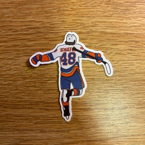 May include: A sticker depicting a hockey player in a white, blue, and orange uniform. The player's jersey displays the number 48 and the name "SCHAEFER". The sticker is placed on a wooden surface.