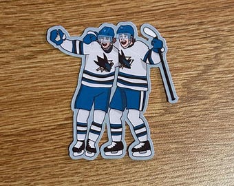 May include: A sticker featuring two cartoon hockey players in white and blue uniforms, celebrating with arms raised. The sticker has a light blue outline and depicts the San Jose Sharks logo.
