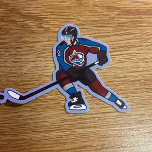 Pode incluir: Um autocolante com um jogador de hóquei em uniforme azul, bordô e branco, com o número 8 visível. O jogador está em ação, com o stick na mão, com o disco à frente. O logótipo da equipa está no peito.