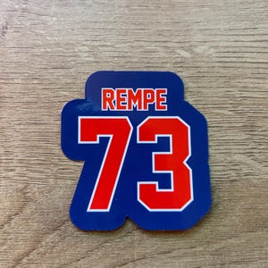 Puede incluir: Una pegatina azul y blanca con la palabra "REMPE" en letras rojas en la parte superior. Debajo, el n&uacute;mero "73" se muestra en rojo con un contorno blanco. La pegatina est&aacute; sobre una superficie de madera.