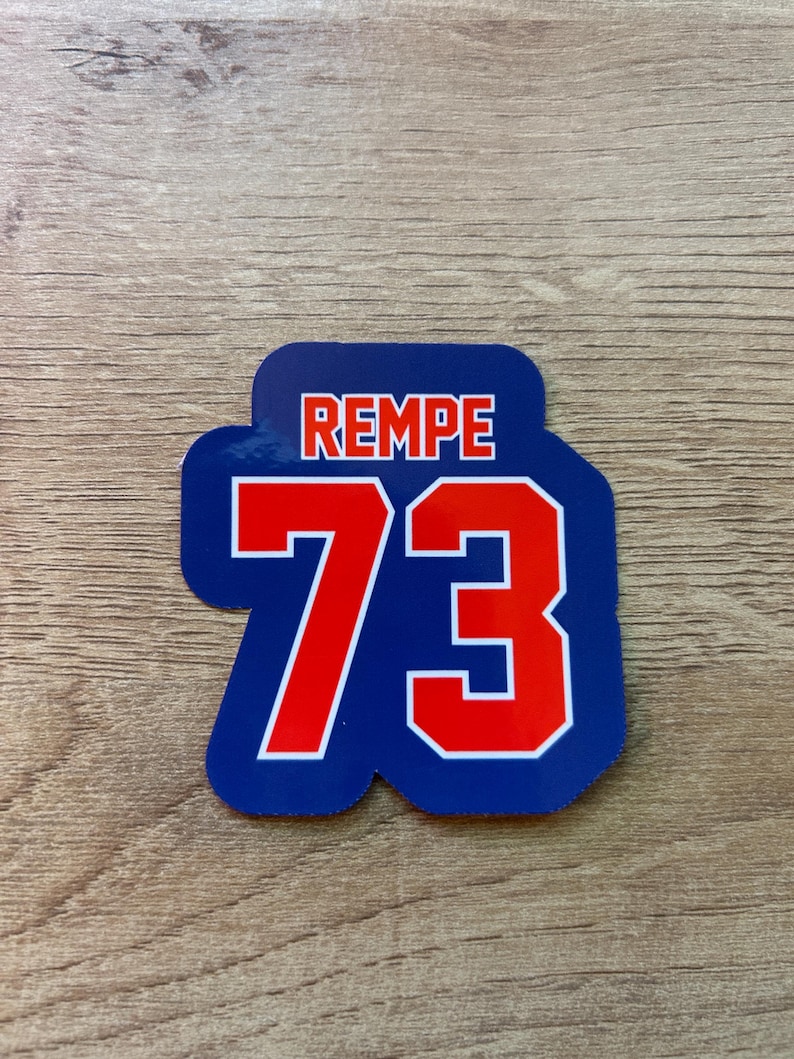 Puede incluir: Una pegatina azul y blanca con la palabra "REMPE" en letras rojas en la parte superior. Debajo, el n&uacute;mero "73" se muestra en rojo con un contorno blanco. La pegatina est&aacute; sobre una superficie de madera.