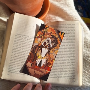 Peut inclure: Un marque-page illustrant une personne en robe blanche et armure noire debout devant un dragon de feu, dans un style de vitrail. Le marque-page est rectangulaire et présente une palette de couleurs brunes, orange et jaunes.