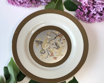 Könnte beinhalten: Ein weißer Keramikteller mit goldfarbenem Rand und einem zentralen Design aus zwei goldenen Vögeln auf einem Zweig mit Blumen. Der Teller ist von grünen Blättern und lila Blüten umgeben.