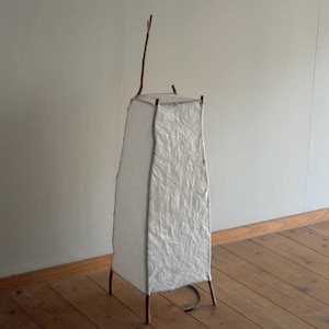 May include: A white paper lampshade with a natural wood branch extending from the top. The lampshade is supported by a black metal frame and sits on a wooden floor.