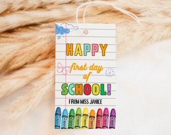 Peut inclure: Une étiquette cadeau rectangulaire avec le texte "HAPPY first day of SCHOOL!" en lettres colorées. L'étiquette comprend une rangée d'illustrations de crayons et le texte "FROM MISS JANICE". L'étiquette est blanche avec une ficelle attachée en haut.