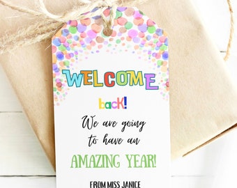 Peut inclure: Une étiquette cadeau avec les mots "WELCOME back! We are going to have an AMAZING YEAR!" en texte coloré. L'étiquette est attachée à un cadeau emballé avec de la ficelle. L'étiquette est signée "FROM MISS JANICE".