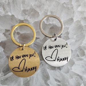 Op de afbeelding: Twee ronde metalen sleutelhangers met ringen. Eén is goudkleurig en de andere is zilverkleurig. Beide zijn voorzien van de gegraveerde zwarte tekst "Ik hou van jou!" gevolgd door een handgetekend hartje en de naam "Harry" in een handschrift.