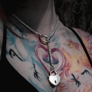 May include: A silver chain choker with a heart-shaped padlock hanging from it. The chain is connected to a ring at the top and the padlock has a keyhole. The choker is worn by a person with tattoos on their chest.