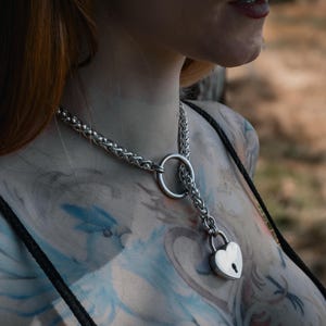 Può includere: Un collare choker a catena argentata con un ciondolo a forma di lucchetto a forma di cuore. La catena è intrecciata e ha un anello grande sul retro del collo.