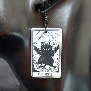 May include: Black and white tarot card earrings featuring a black cat with wings and a halo. The card reads "The Devil".
