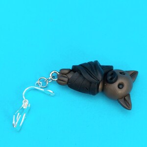May include: A brown and black bat charm with a silver clip-on earring finding. The bat is hanging upside down with its wings folded.