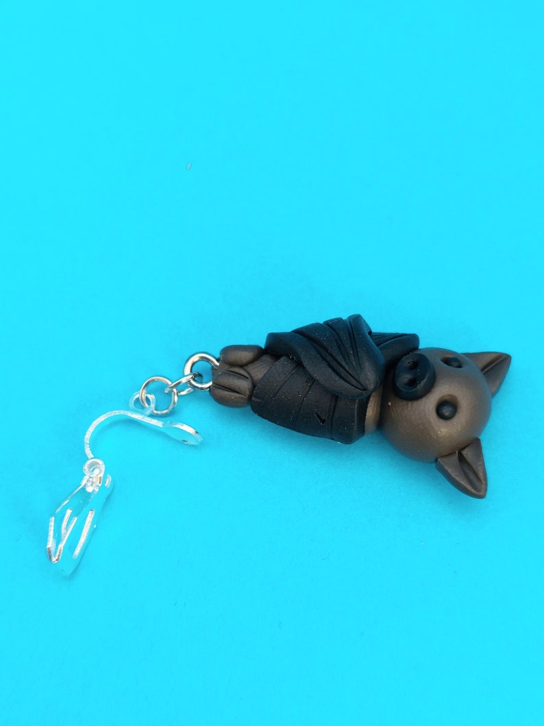May include: A brown and black bat charm with a silver clip-on earring finding. The bat is hanging upside down with its wings folded.