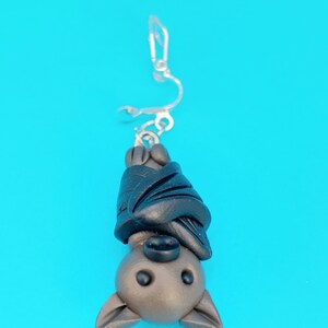May include: A brown and black bat charm hanging from a silver clip-on earring. The bat is upside down with its wings wrapped around its body.