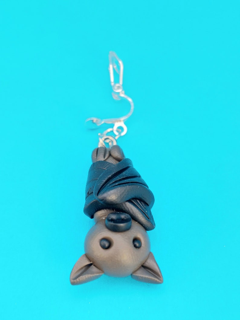 May include: A brown and black bat charm hanging from a silver clip-on earring. The bat is upside down with its wings wrapped around its body.